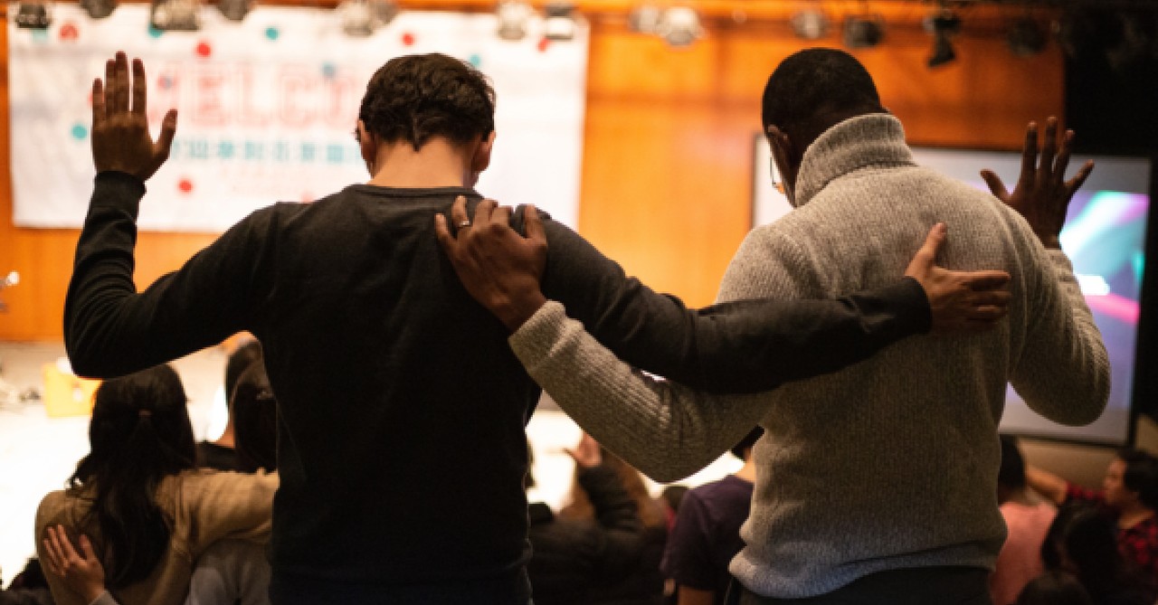 Two men with arms around each other and one arm raised, praying together